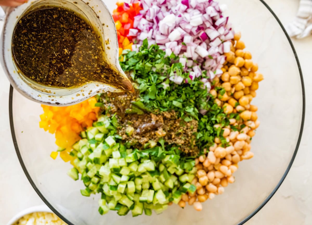 Dressing being poured from a small pitcher into a glass mixing bowl of beans, diced vegetables, and herbs.
