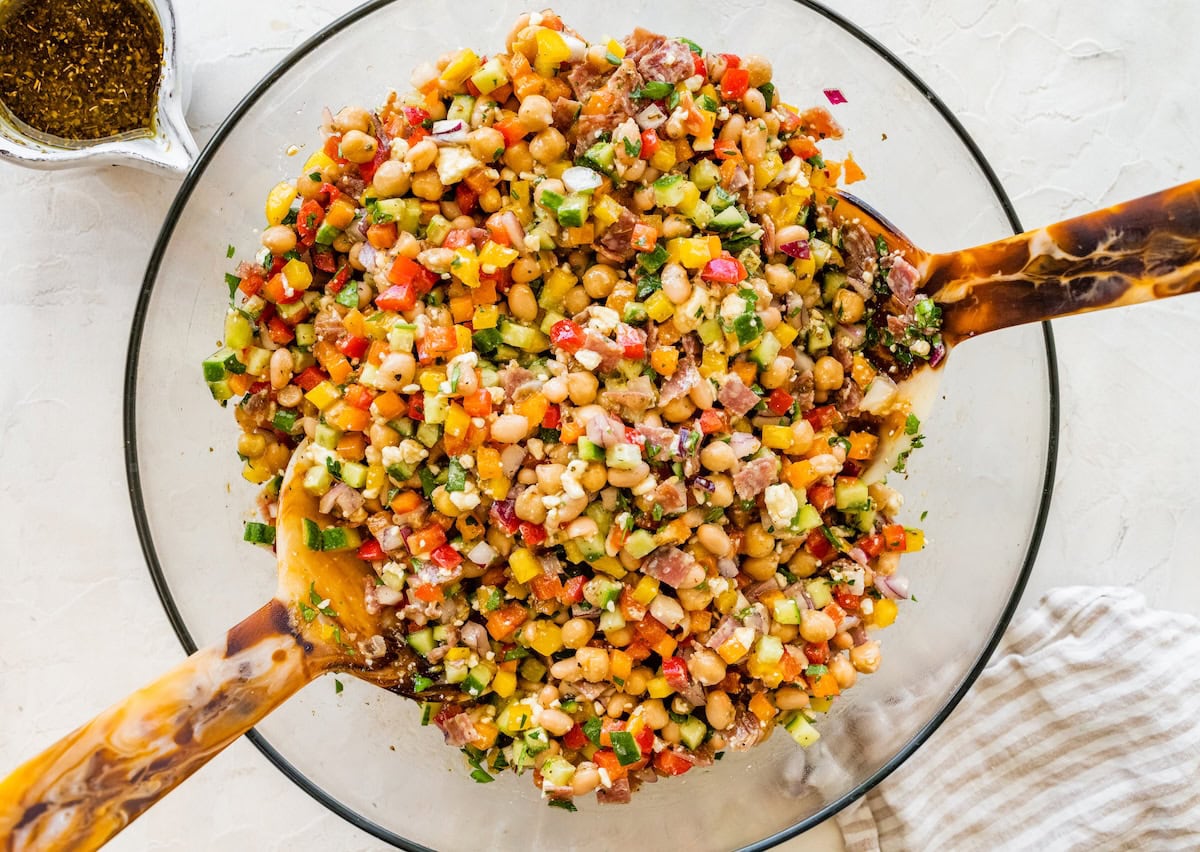 Overhead view of fully mixed bean salad with diced vegetables, beans, cheese, and dressing in a large glass mixing bowl.