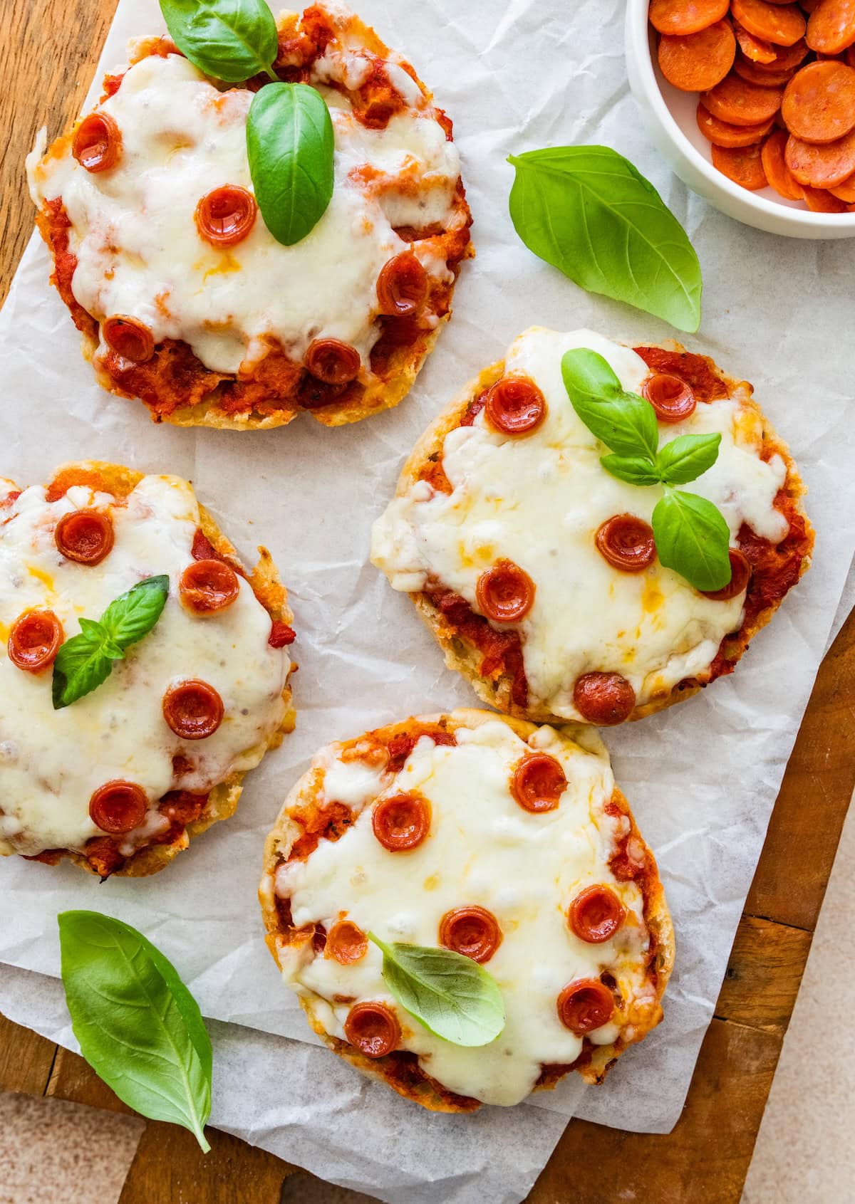 English muffin pizzas topped with melted cheese, mini pepperoni and basil leaves, served on parchment.