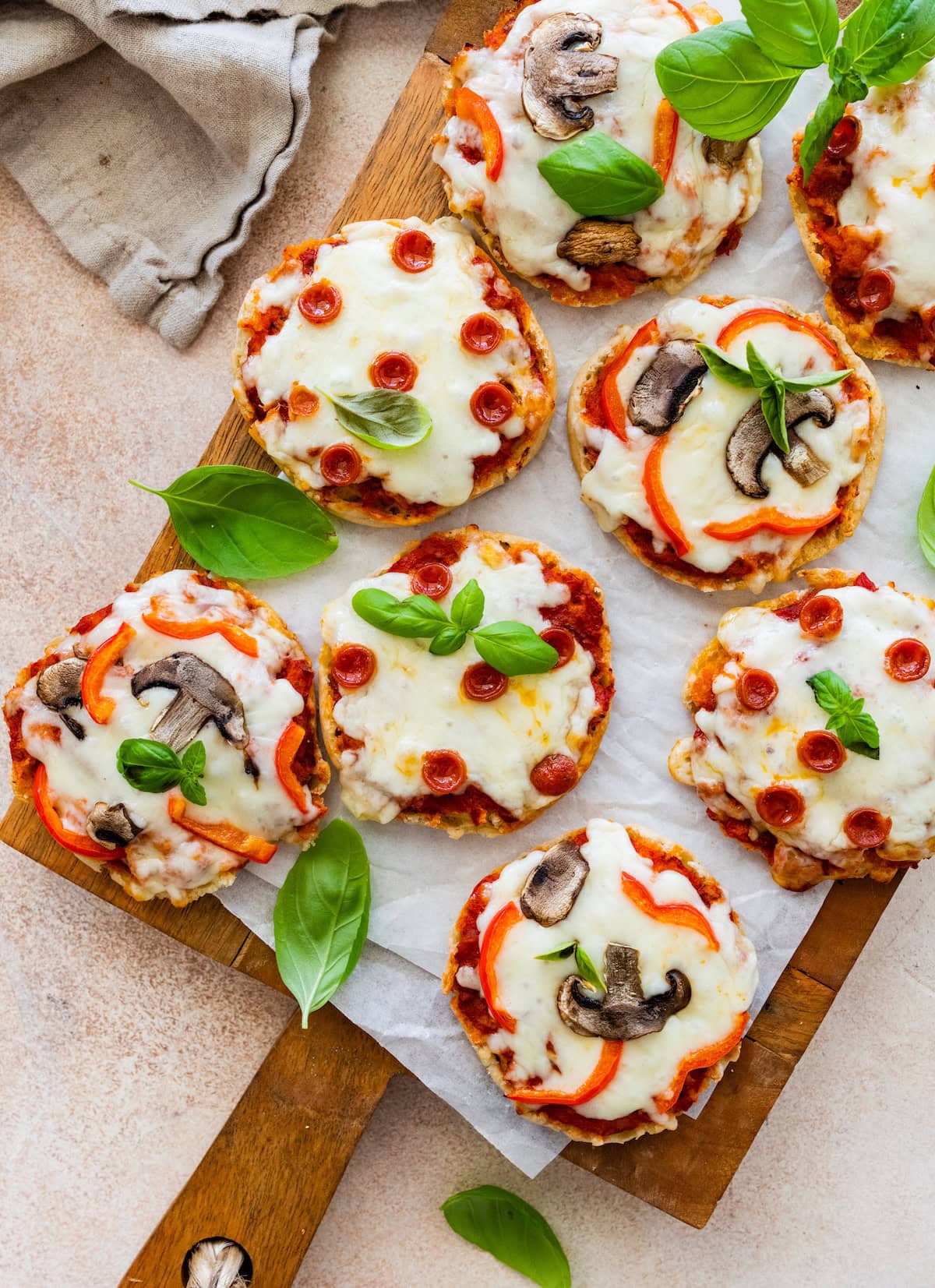 English muffin pizzas with pepperoni and mushrooms on a wooden board, topped with basil.