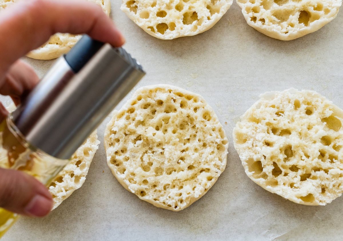 Spraying English muffin halves with olive oil before adding toppings.