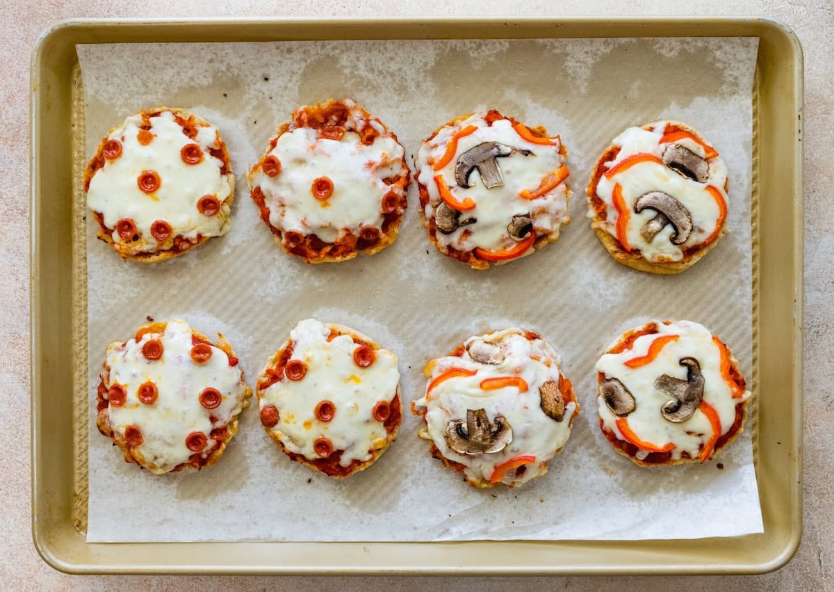 Baked English muffin pizzas on a baking sheet with melted mozzarella and toppings.