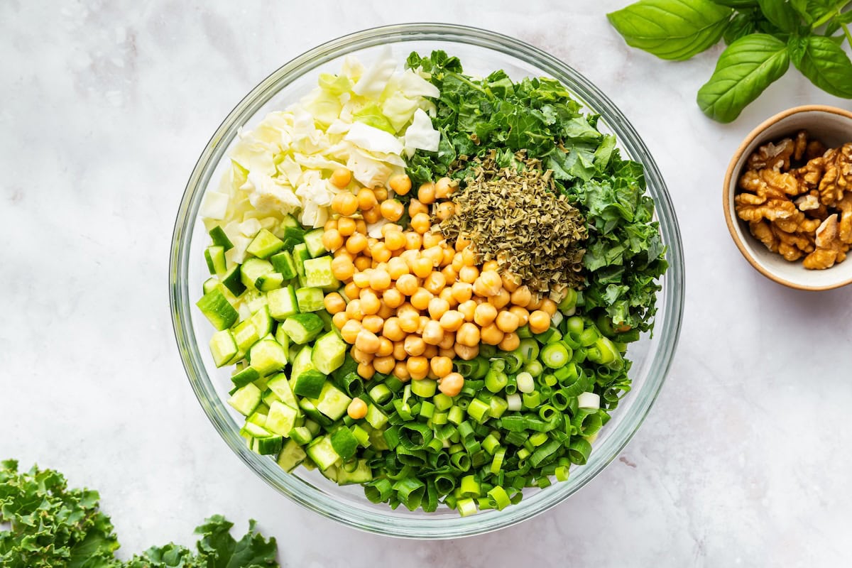 Unmixed green goddess salad ingredients in a bowl, including chopped kale, cabbage, cucumber, green onion, chickpeas, and dried herbs.