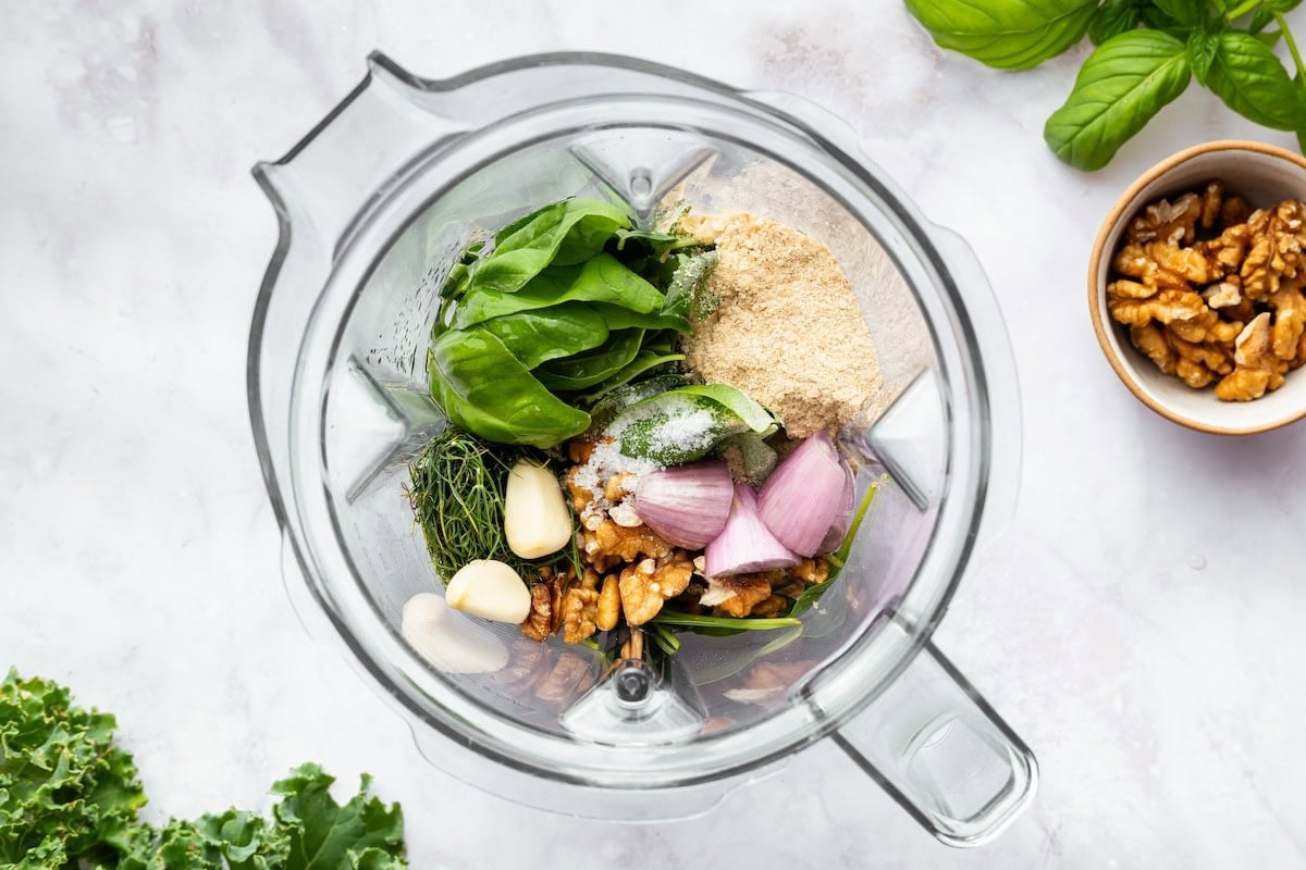 Blender filled with ingredients for green goddess dressing, including spinach, basil, shallot, garlic, nutritional yeast, and walnuts.