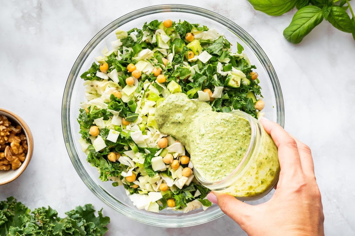 Hand pouring creamy green goddess dressing over chopped salad with chickpeas, kale, and cabbage in a glass bowl.