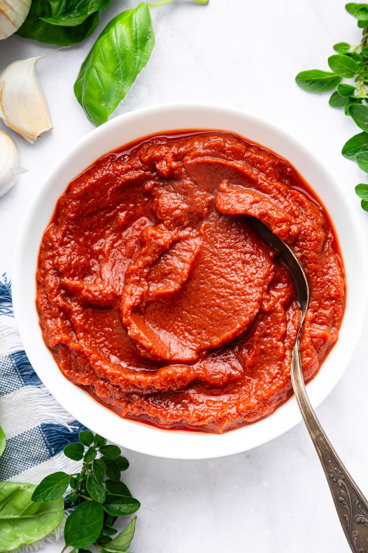Bowl of thick homemade pizza sauce with a spoon, surrounded by fresh herbs.