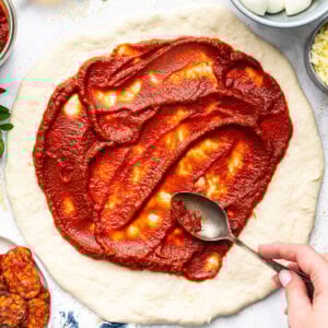 Spreading homemade pizza sauce over pizza dough with a spoon, ready for toppings.