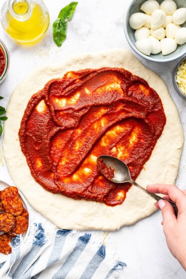Spreading homemade pizza sauce over pizza dough with a spoon, ready for toppings.