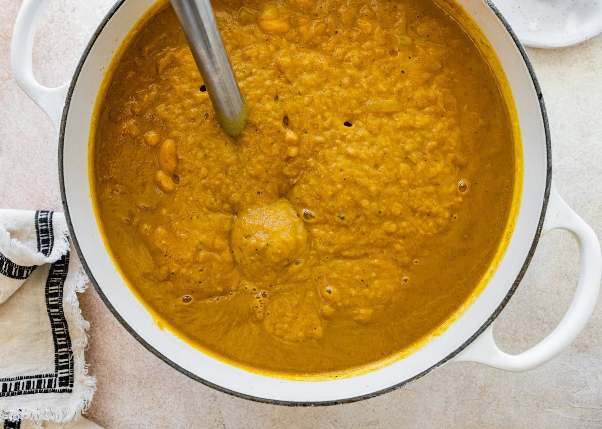 An immersion blender used to blend a pumpkin curry soup in a large pot.