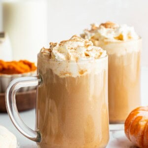 Two glass mugs filled with pumpkin spice lattes topped with whipped cream and cinnamon, set among pumpkins, milk, and fall spices.