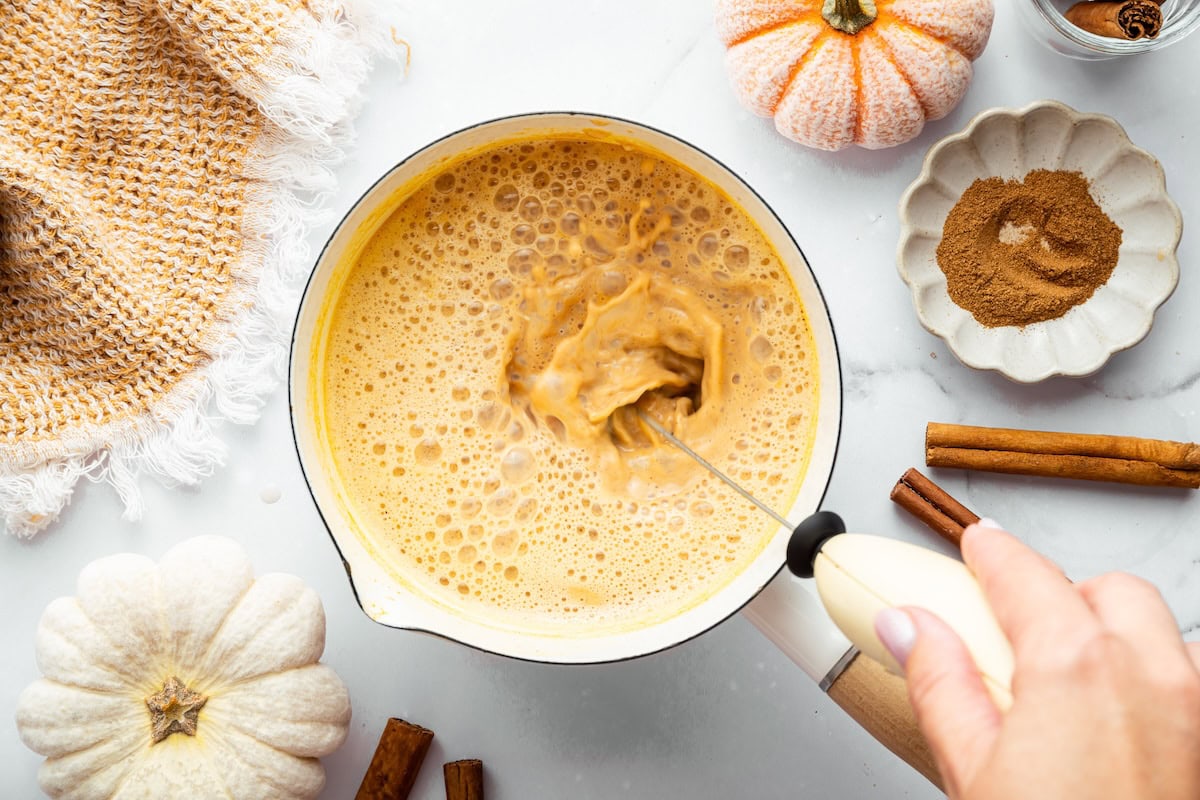 Pumpkin spice latte mixture being frothed in a saucepan with a handheld frother, surrounded by fall spices and pumpkins.