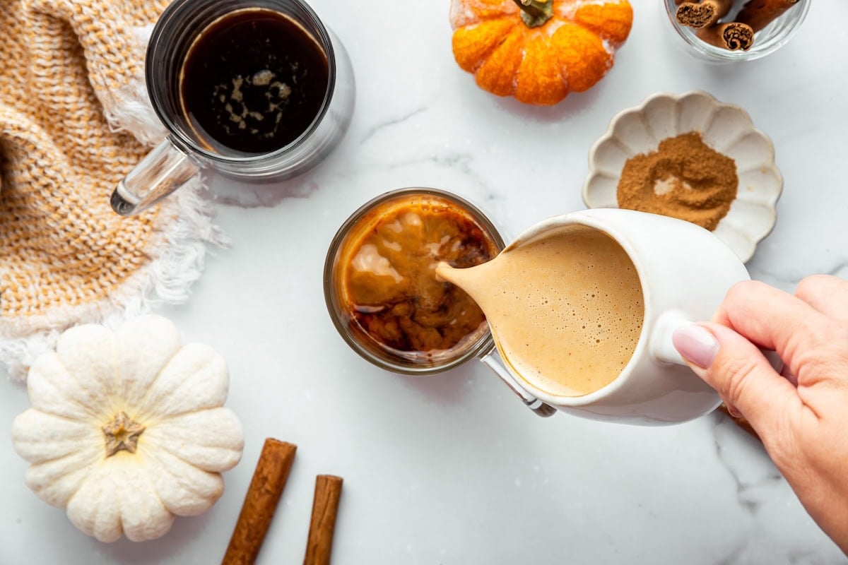 Pumpkin spice latte mixture being poured from a small pitcher into a glass of coffee, with pumpkins and cinnamon sticks on the side.