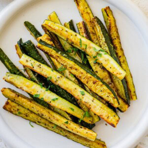 Golden roasted zucchini sticks on a white plate, garnished with chopped parsley.