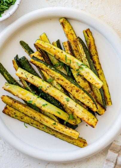 Golden roasted zucchini sticks on a white plate, garnished with chopped parsley.