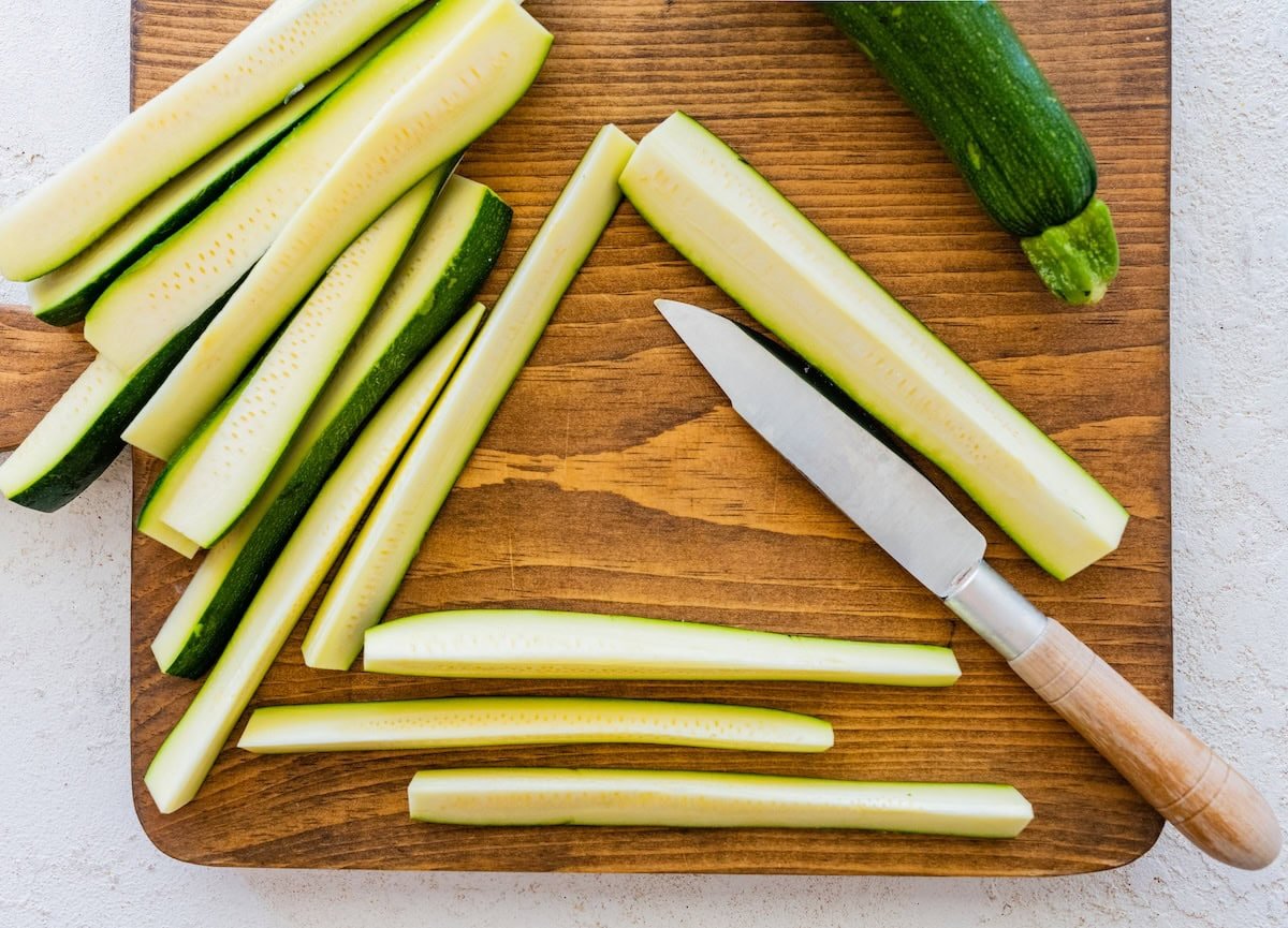 Fresh zucchini sliced into sticks on a wooden cutting board with a knife.