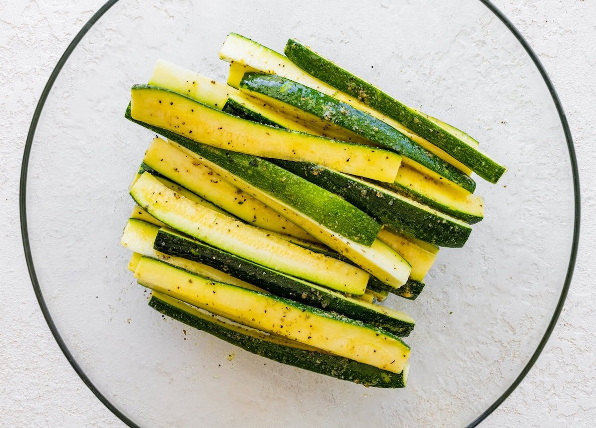 Zucchini sticks tossed with olive oil and seasonings in a glass mixing bowl.