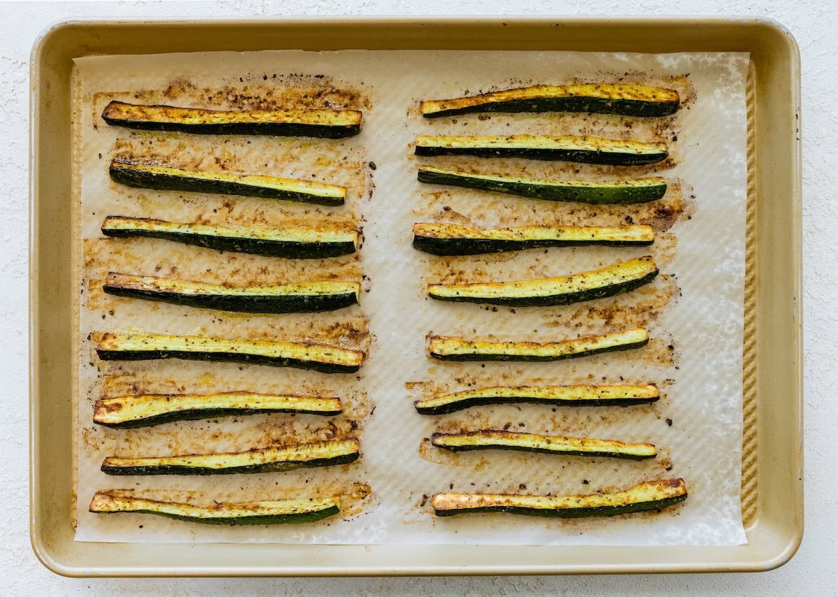 Roasted zucchini sticks on a parchment-lined baking sheet, golden and crispy with browned edges.