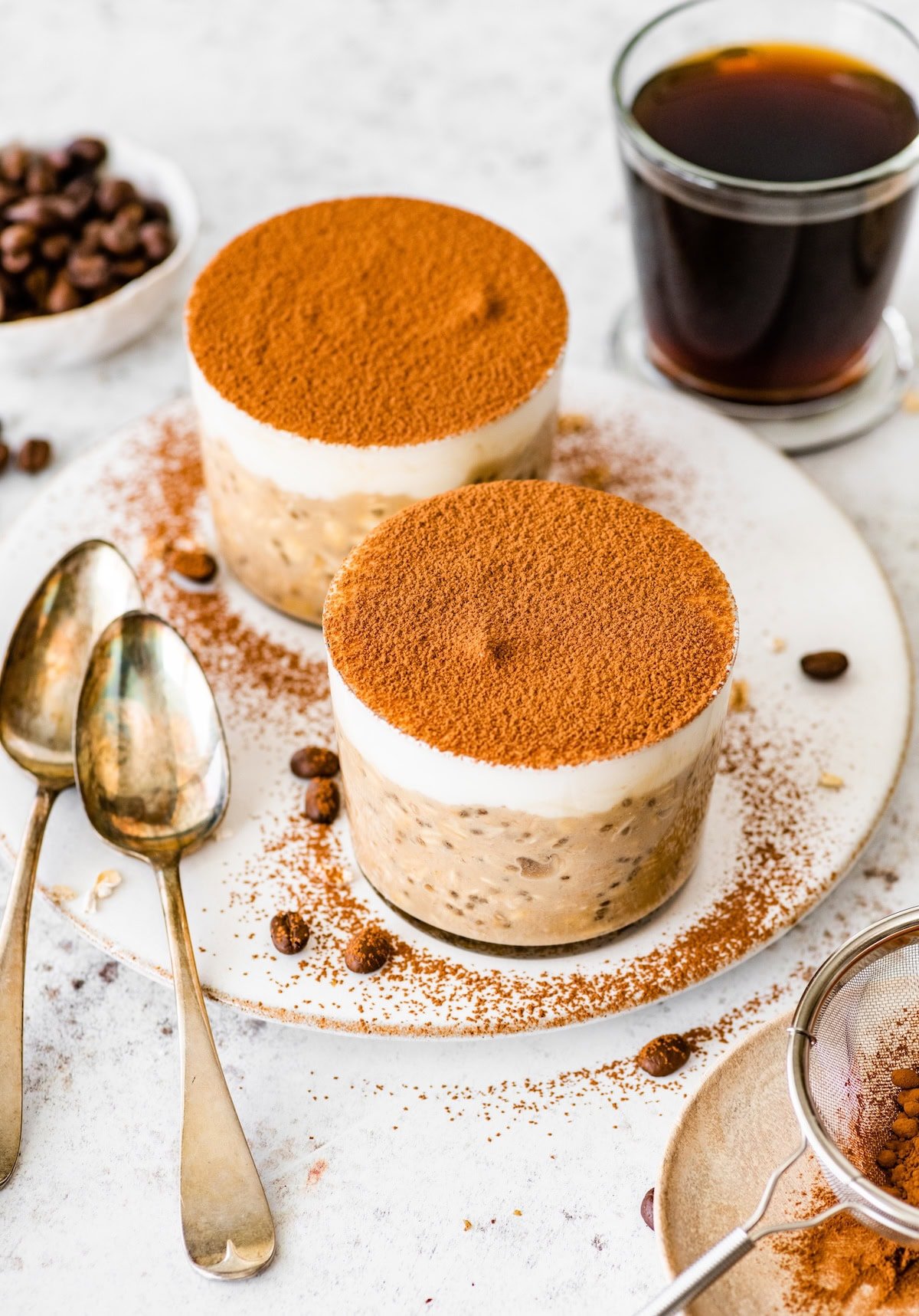 Two jars of tiramisu overnight oats topped with cocoa powder, served with coffee and vintage spoons.