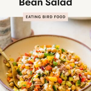 Colorful bean salad with chopped vegetables and feta in a beige bowl with a gold spoon.