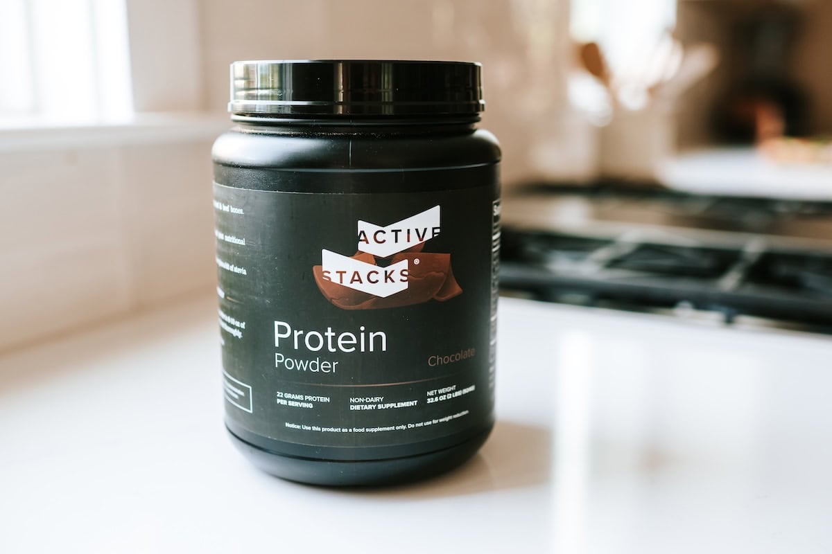 Black container of Active Stacks chocolate protein powder on a white kitchen counter, labeled with 22 grams of protein per serving.