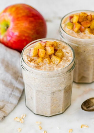 Two jars of apple cinnamon overnight oats topped with diced apples, with a fresh apple behind the jars.