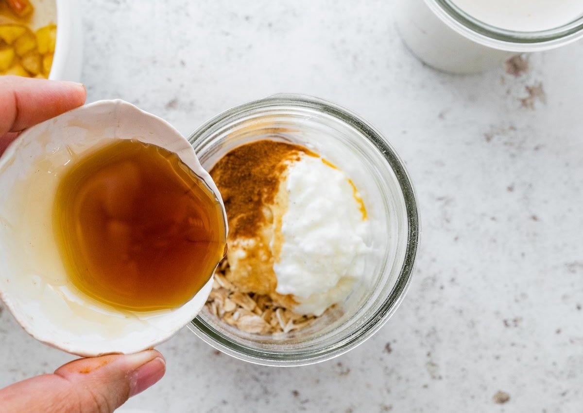 Hand pouring maple syrup into a glass jar containing oats, yogurt, and cinnamon.