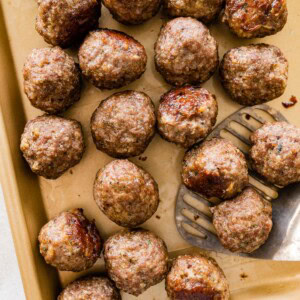 Baked meatballs resting on a parchment-lined sheet pan with browned tops and a metal spatula taking a few from the pan.