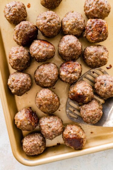 Baked meatballs resting on a parchment-lined sheet pan with browned tops and a metal spatula taking a few from the pan.