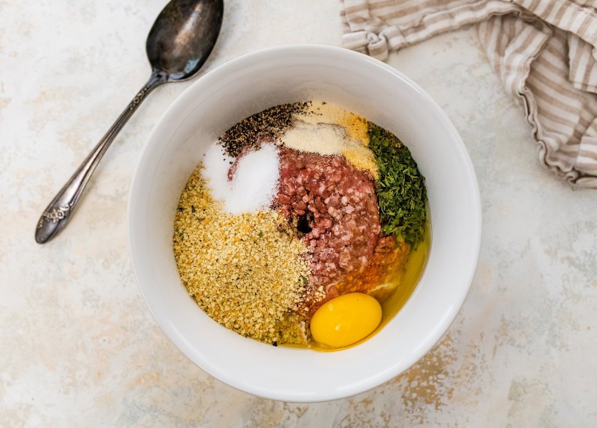 Mixing bowl with ground beef, breadcrumbs, egg, parsley, and seasonings before combining.