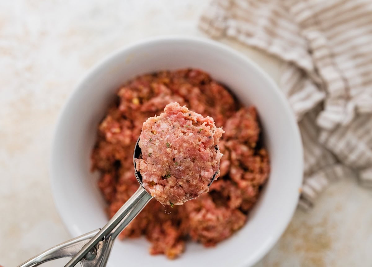 Scoop of raw meatball mixture held above a bowl of prepared mixture.