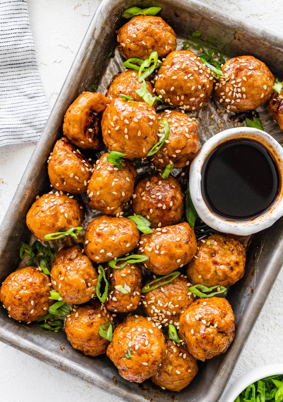 Baked BBQ chicken meatballs topped with sesame seeds and green onions, served in a metal tray with a small bowl of Japanese BBQ sauce.