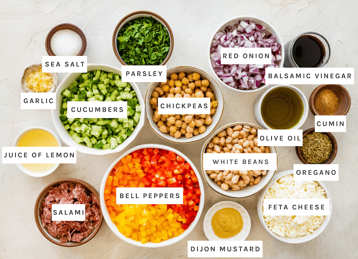 Dense Bean Salad ingredients: Sea salt, parsley, red onion, balsamic vinegar, garlic, cucumbers, chickpeas, olive oil, cumin, juice of lemon, drained beans, oregano, bell peppers, feta cheese, salami, Dijon mustard