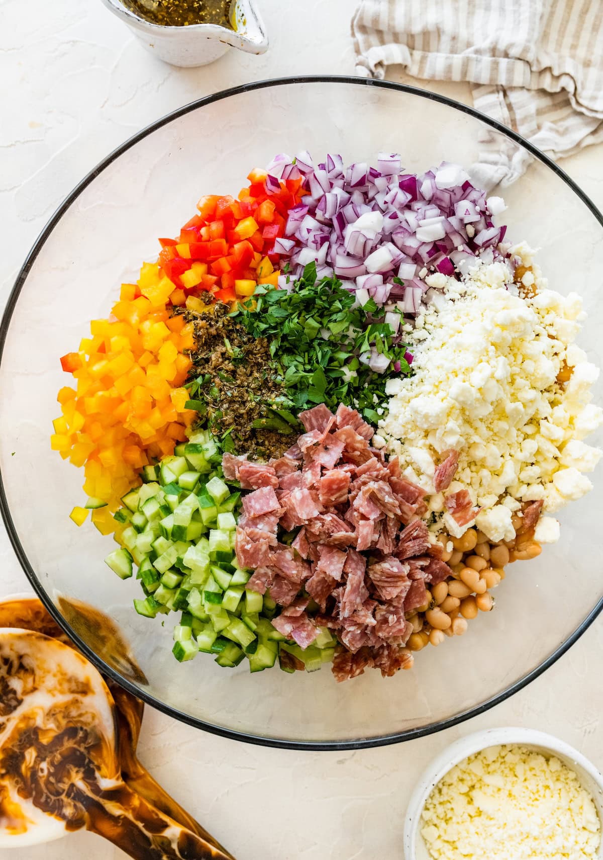 Chopped cucumbers, bell peppers, red onion, parsley, feta, beans, and salami in a mixing bowl for dense bean salad.
