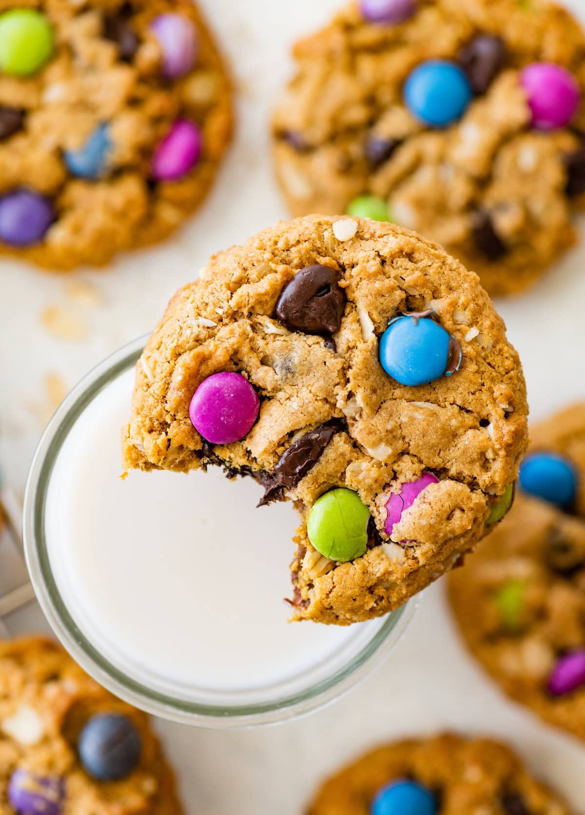 A soft monster cookie with colorful candy pieces and chocolate chips sits on the rim of a glass of milk with a bite missing from the edge.