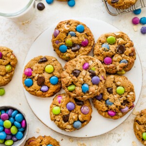 A full plate of monster cookies surrounded by scattered candies, a cooling rack, and two glasses of milk on the table.