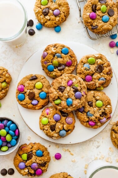 A full plate of monster cookies surrounded by scattered candies, a cooling rack, and two glasses of milk on the table.