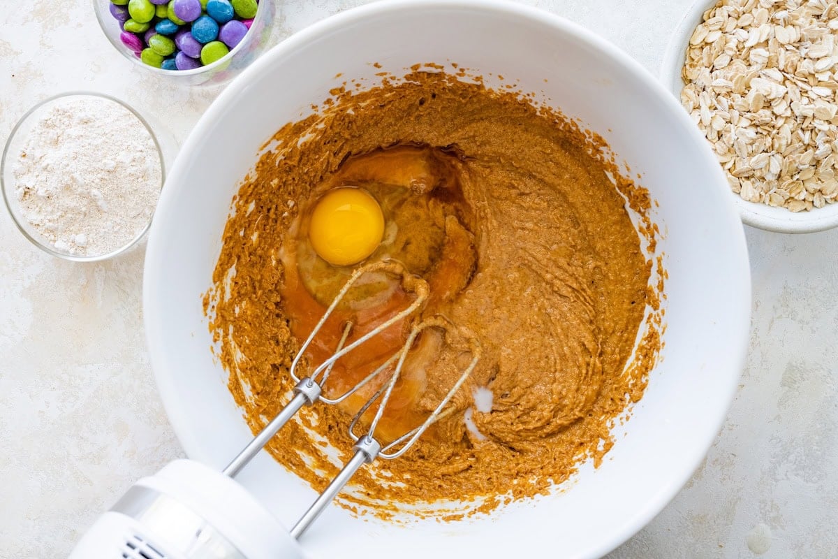 A mixing bowl of monster cookie batter with a cracked egg sitting on top, ready to be blended in with a hand mixer.