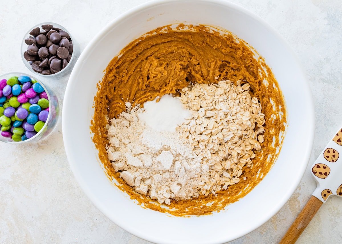 A mixing bowl with cookie batter topped with oats, flour and baking soda before being mixed.