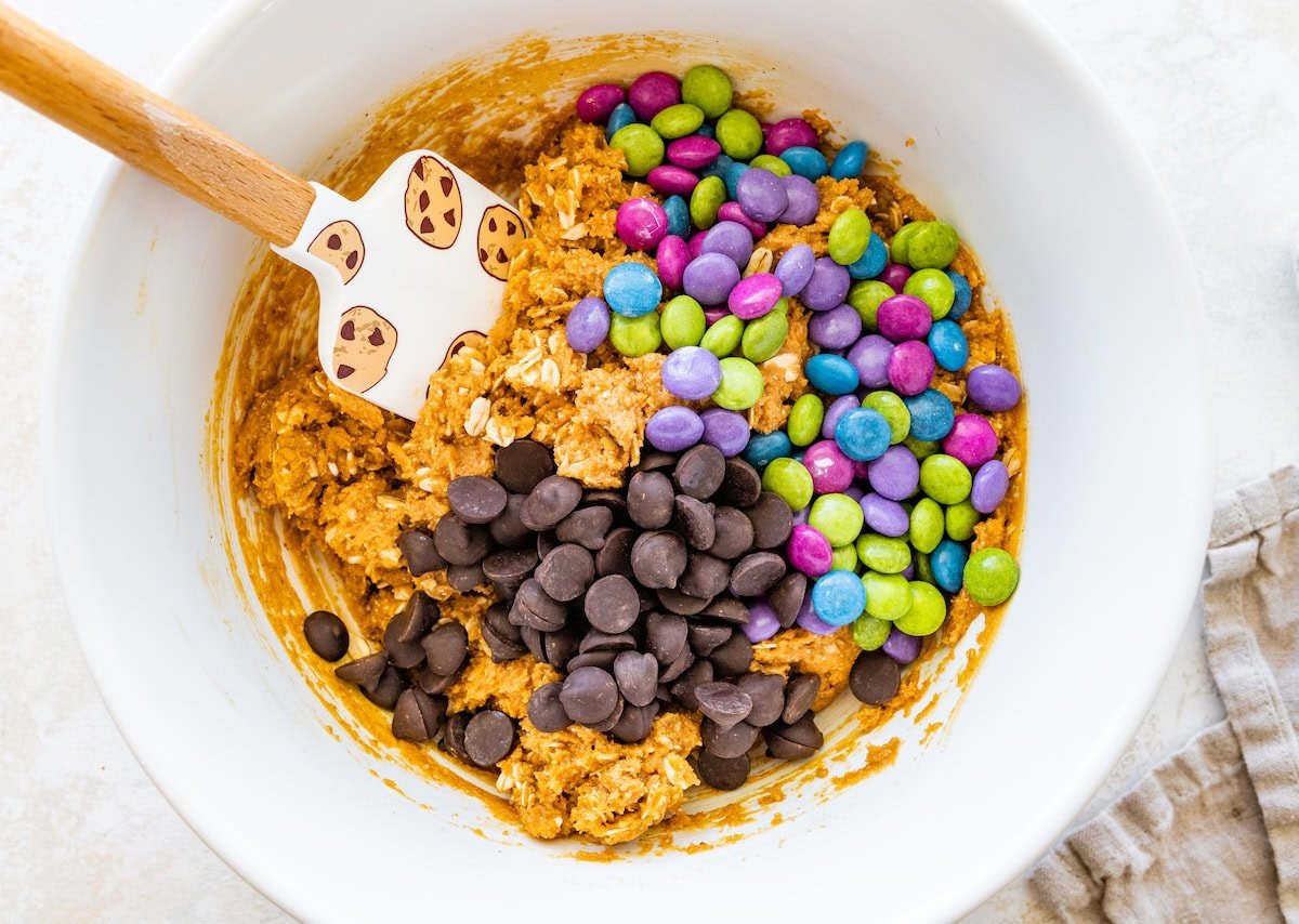 A mixing bowl filled with monster cookie dough topped with a large pile of chocolate chips and bright candy-coated chocolates before stirring.