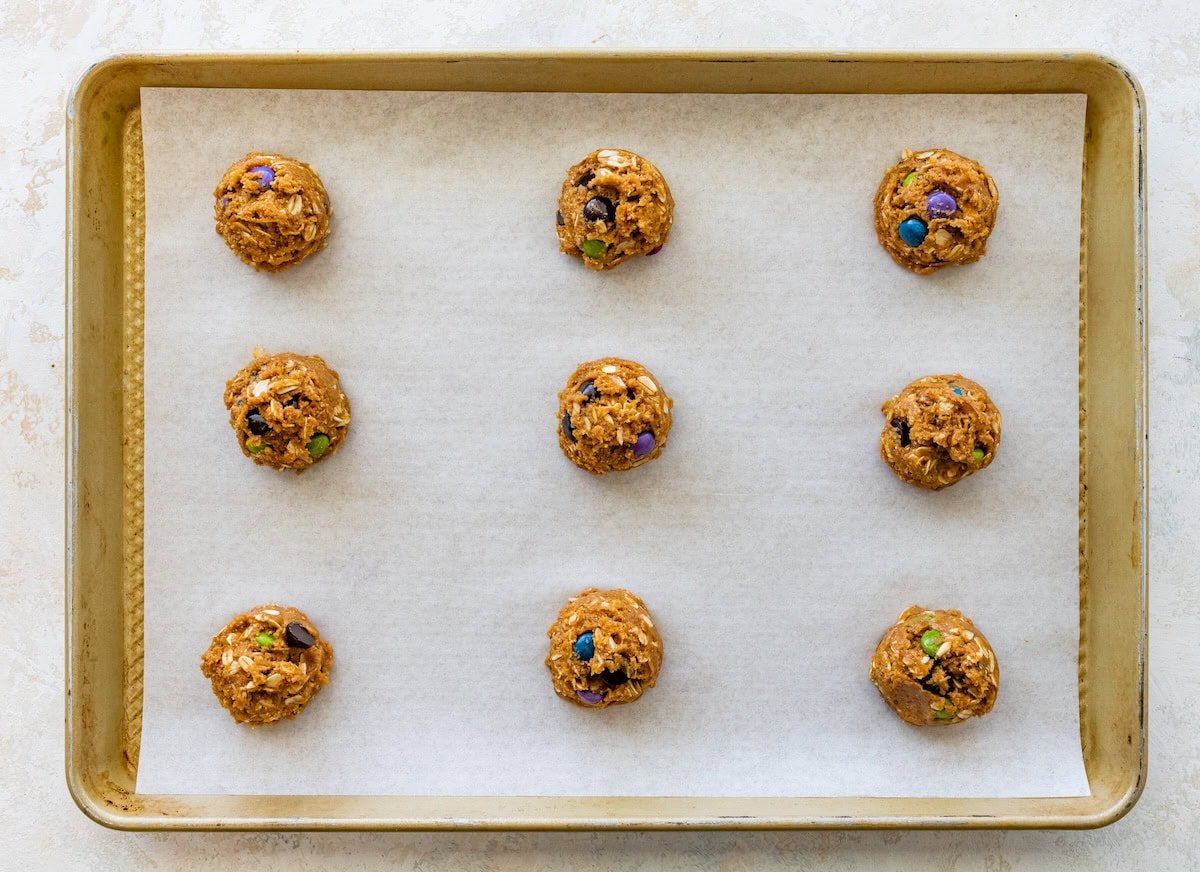 Nine scoops of monster cookie dough spaced evenly on a parchment-lined baking sheet, ready to bake.