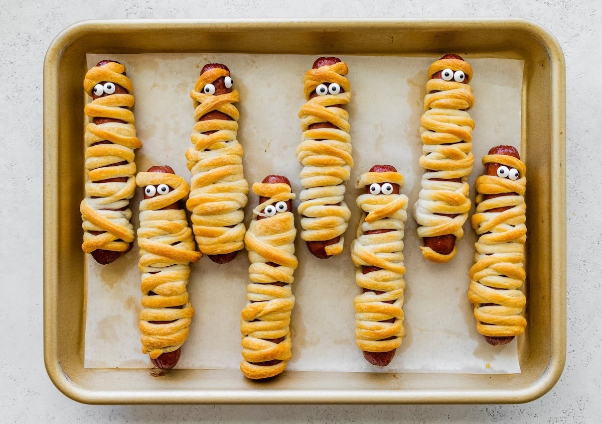 Freshly baked mummy hot dogs with candy eyes on a sheet pan, wrapped in crescent roll dough for Halloween.