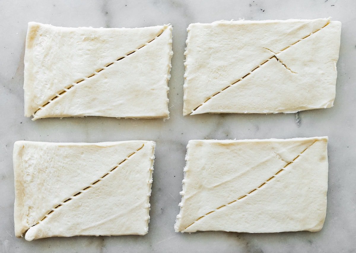 Crescent roll dough cut into rectangles on a marble surface, ready to be sliced into strips for mummy hot dogs.