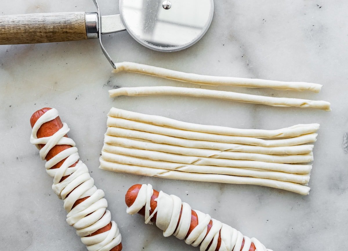 Thin crescent dough strips cut with a pizza cutter, ready to wrap hot dogs for mummy hot dogs.