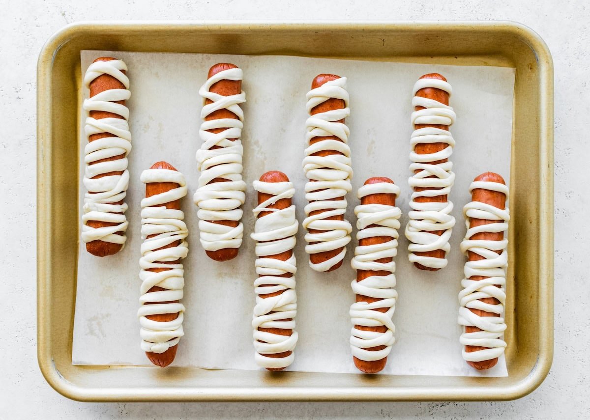 Unbaked mummy hot dogs wrapped in crescent dough strips on a baking sheet before going in the oven.