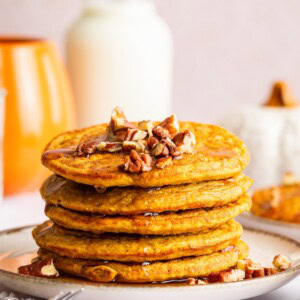 Tall stack of pumpkin protein pancakes on a plate topped with pecans and drizzled with syrup.