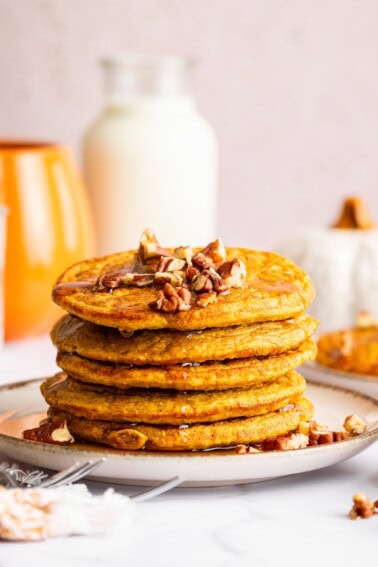 Tall stack of pumpkin protein pancakes on a plate topped with pecans and drizzled with syrup.
