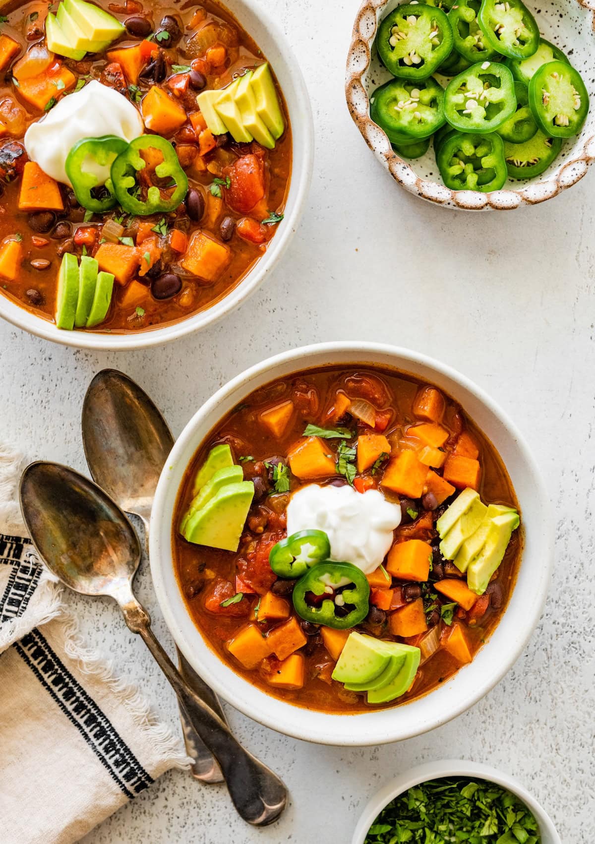 Two bowls of sweet potato black bean chili topped with sour cream, jalapeño slices, and avocado.