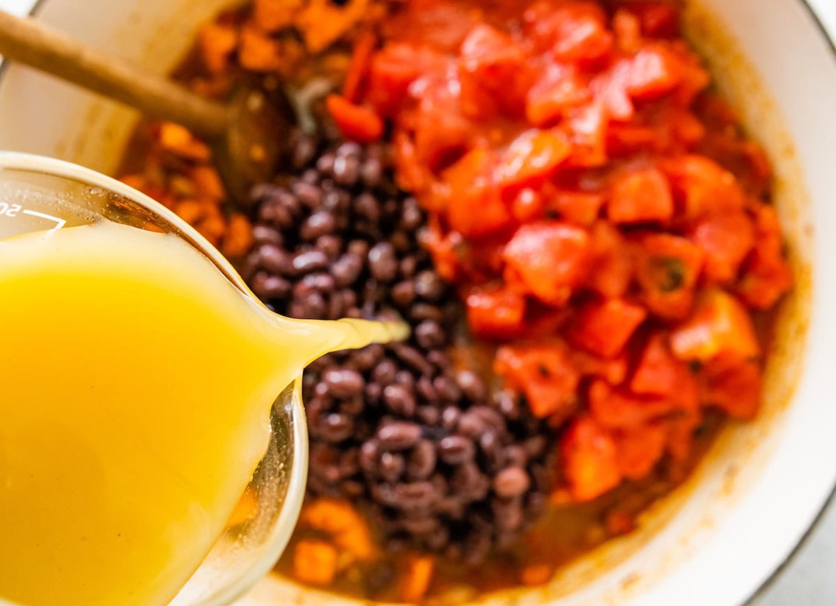 A large pot of beans, tomato, sweet potato, and spices with a broth being poured into the pot.