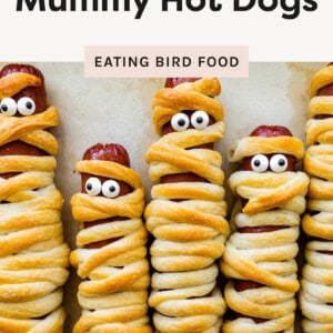 Five mummy hot dogs wrapped in golden crescent roll dough with candy eyes, lined up side by side on parchment paper