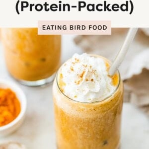 A glass of iced pumpkin spice latte topped with whipped cream and sprinkled with pumpkin spice, with bowls of pumpkin puree and spice in the background.